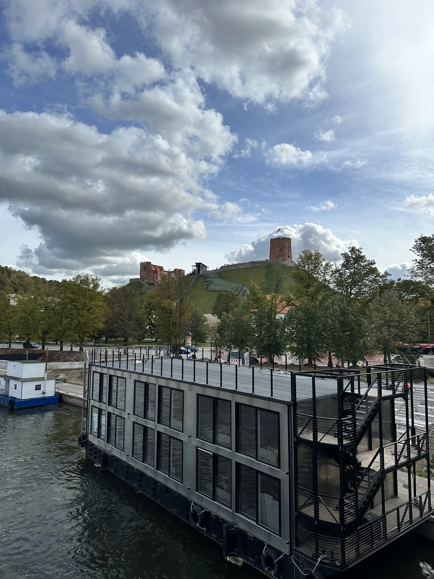 A modern two-story grey houseboat on the Neris River with the historic brick Gediminas' Tower on a green hill under a cloudy sky in the background.