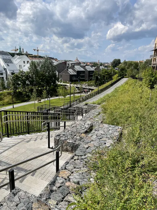A high-angle view of a modern paved pedestrian path and stairs with metal handrails winding down a grassy hill lined with gabion stone walls, overlooking contemporary residential buildings in Vilnius.