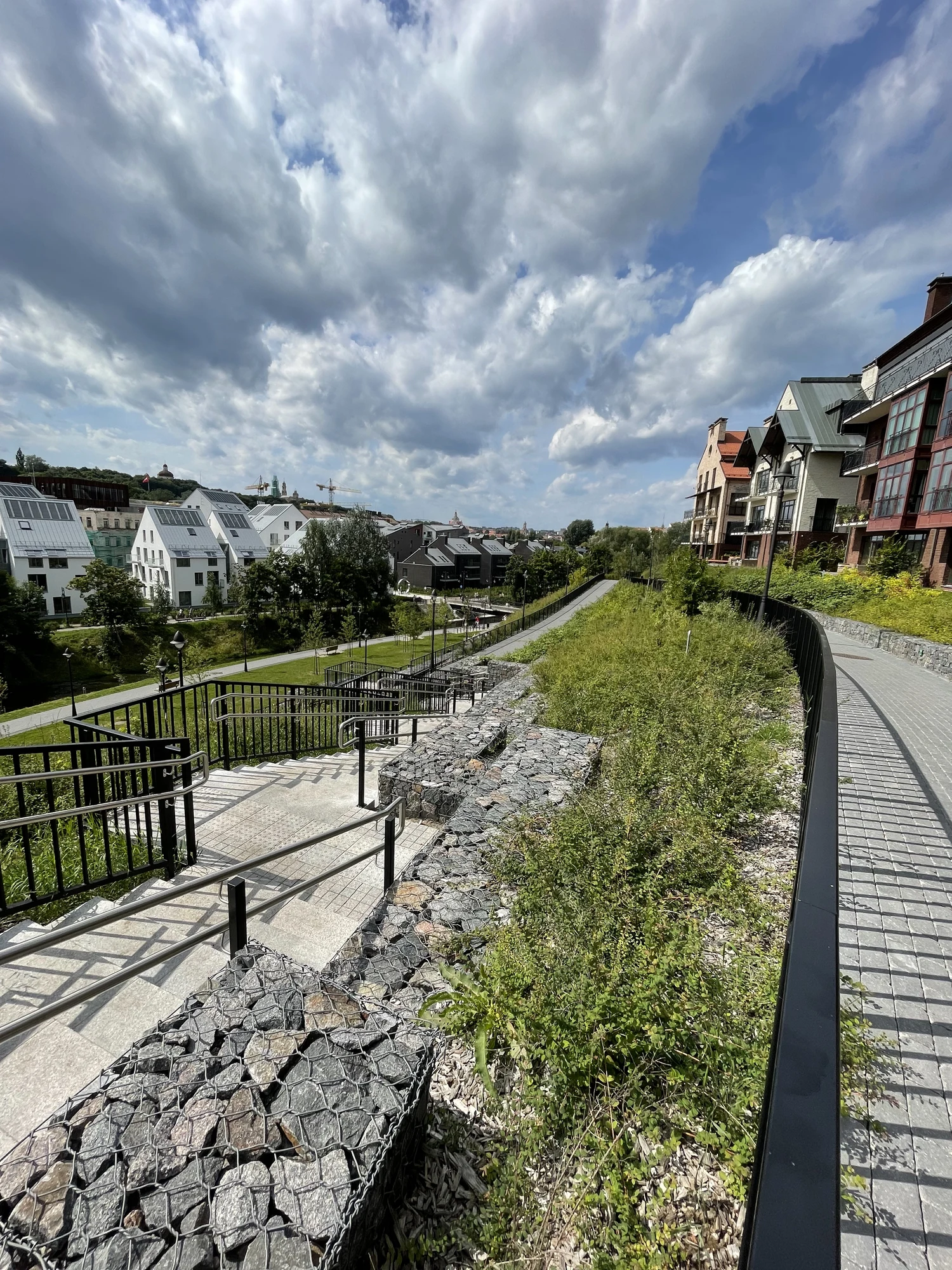 A high-angle view of a modern landscaped hillside in Vilnius, featuring stone gabion walls, paved stairs, and pathways alongside contemporary residential buildings under a cloudy sky.