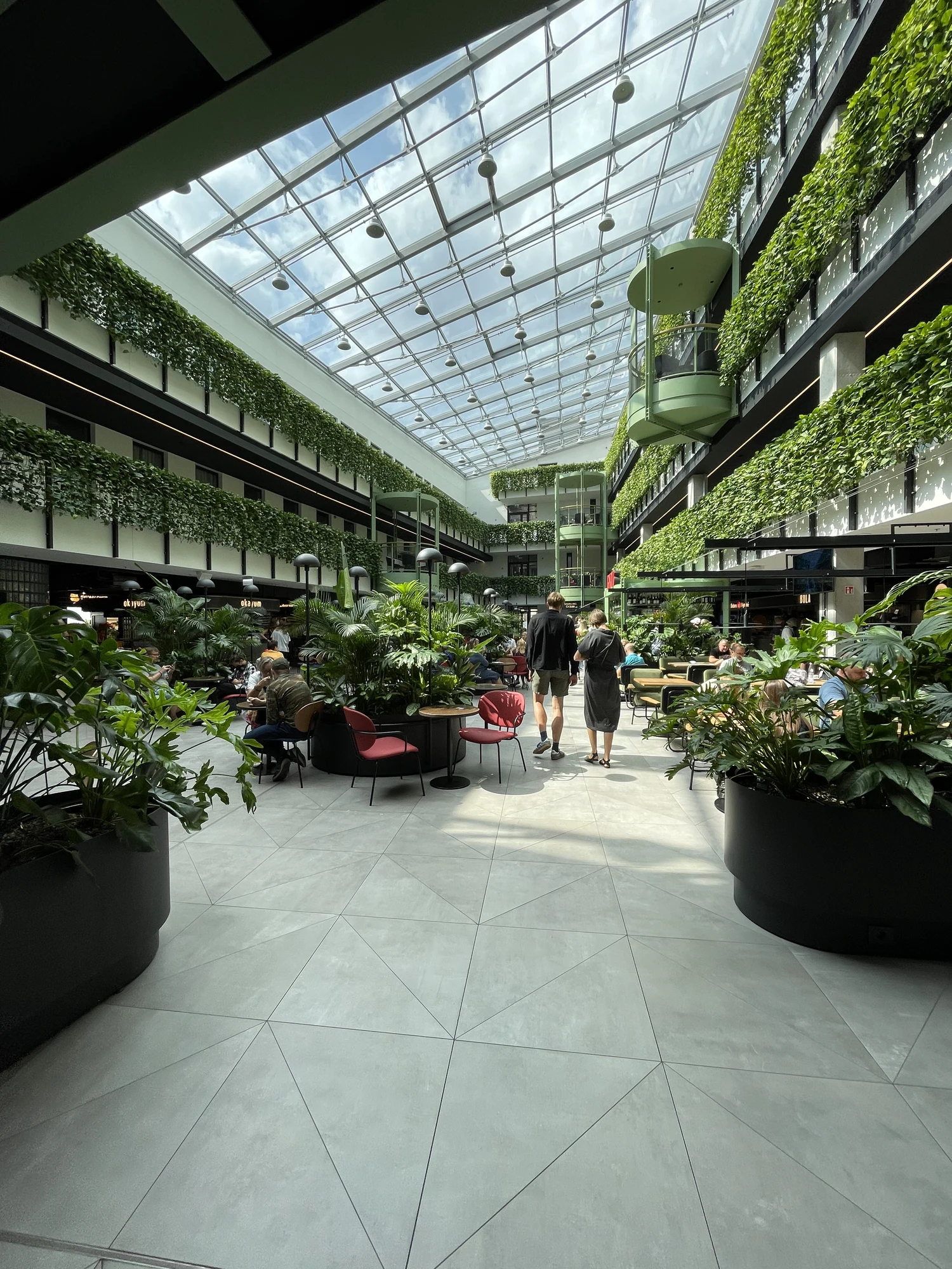 A bright, multi-story indoor atrium with a large glass skylight ceiling, filled with lush green plants, hanging vines, and people sitting at tables in a modern food market.