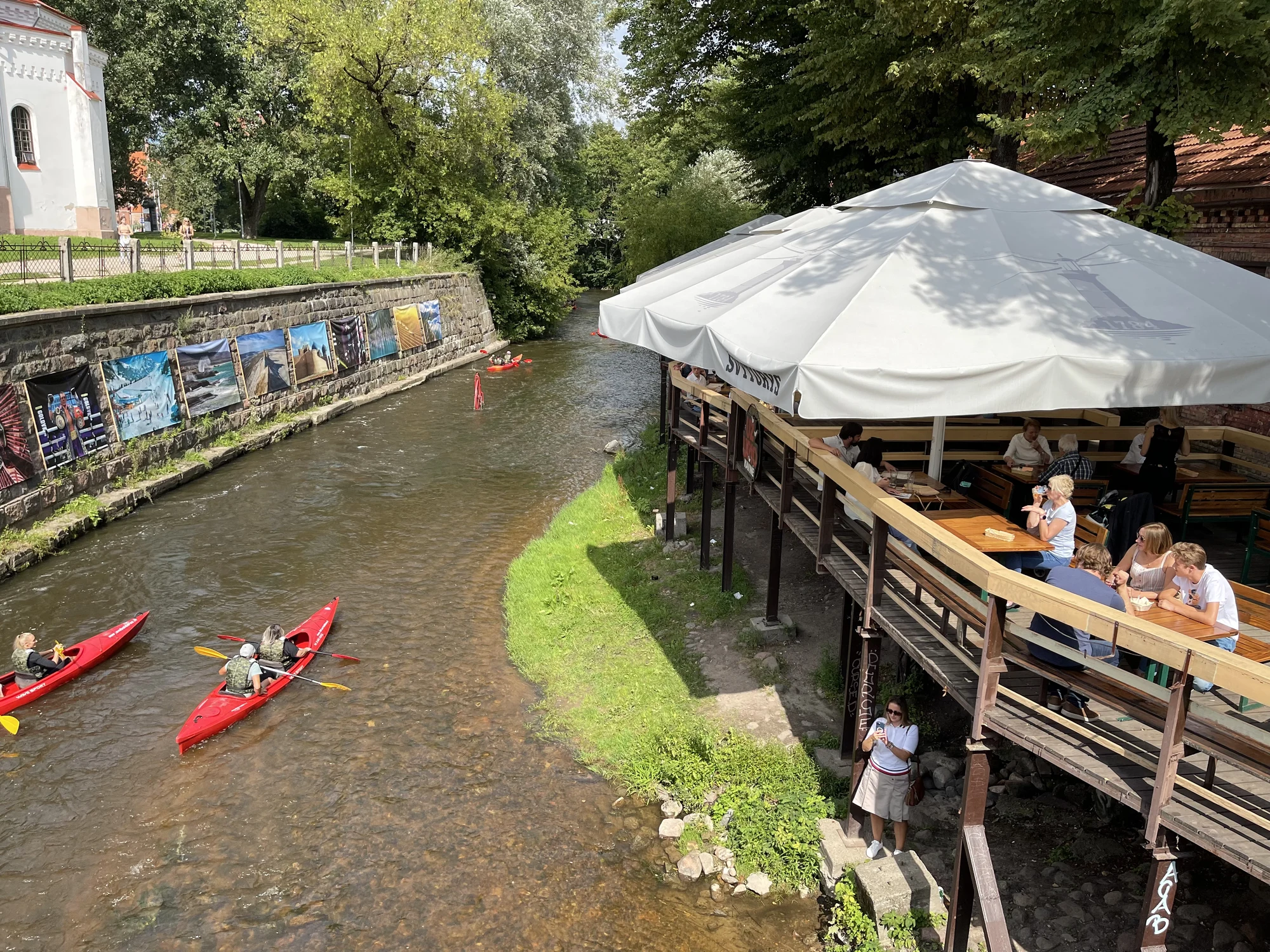 High-angle view of red kayaks navigating the shallow Vilnelė River next to an outdoor cafe terrace with large white umbrellas in Vilnius.