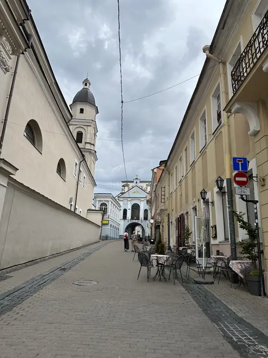 A cobblestone street in Vilnius Old Town lined with historic pastel-colored buildings and outdoor cafe tables, leading towards the Gates of Dawn under a cloudy sky.