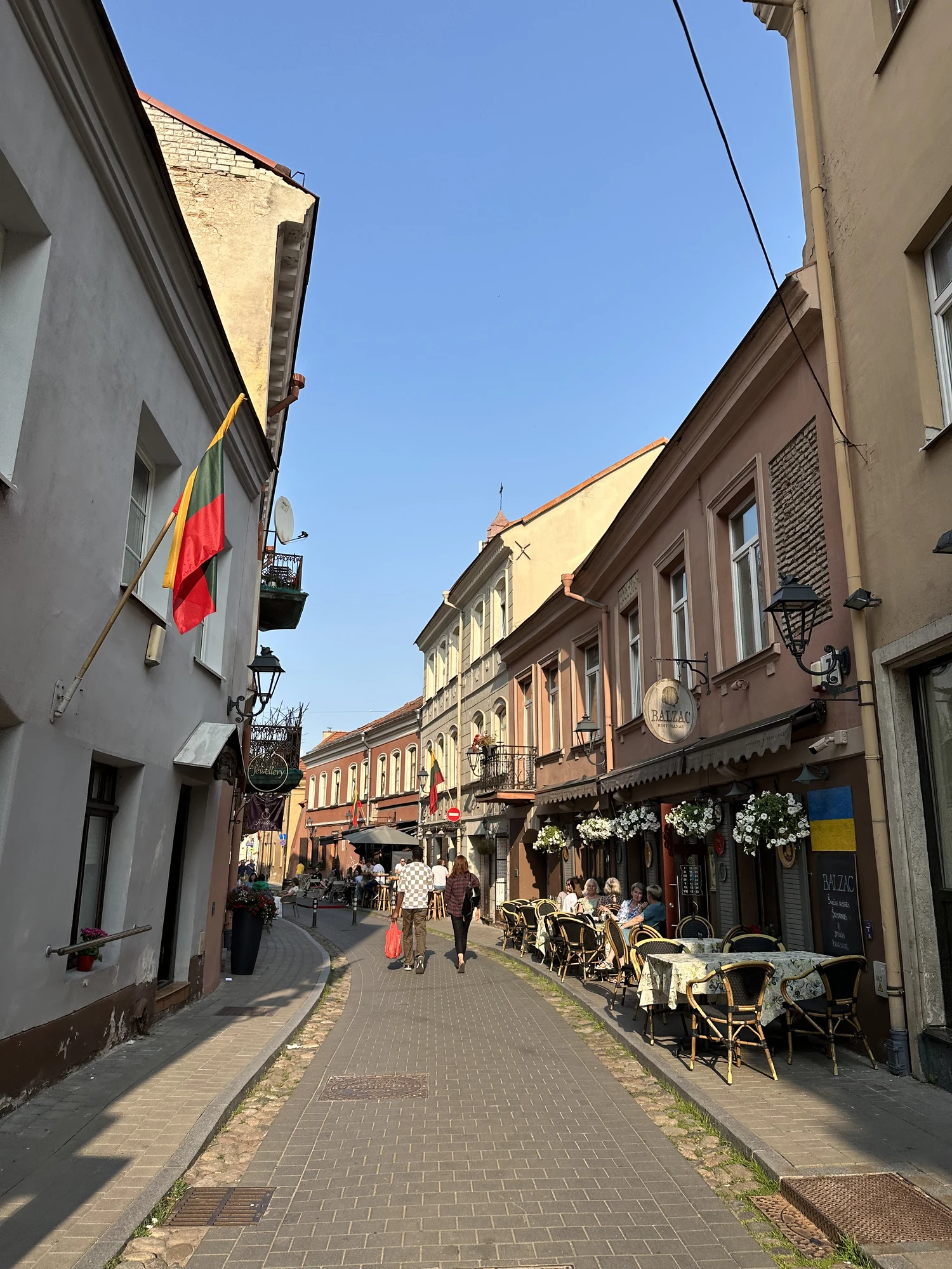 A narrow paved street in Vilnius Old Town lined with historic pastel-colored buildings, outdoor cafe seating, and people walking under a clear blue sky.