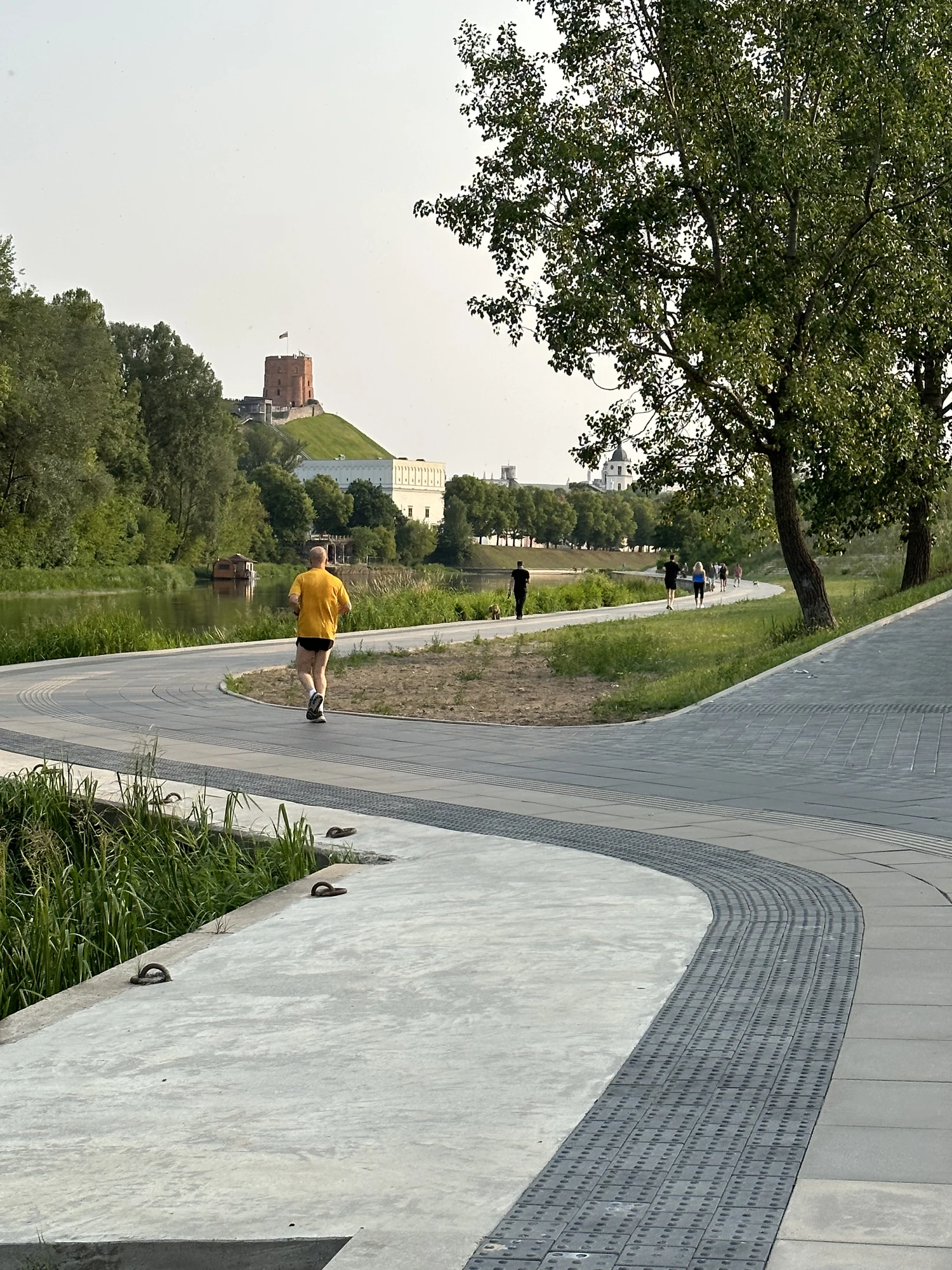 A man in a yellow shirt jogs on a paved riverside path in Vilnius, with Gediminas' Tower visible on a green hill in the distance.