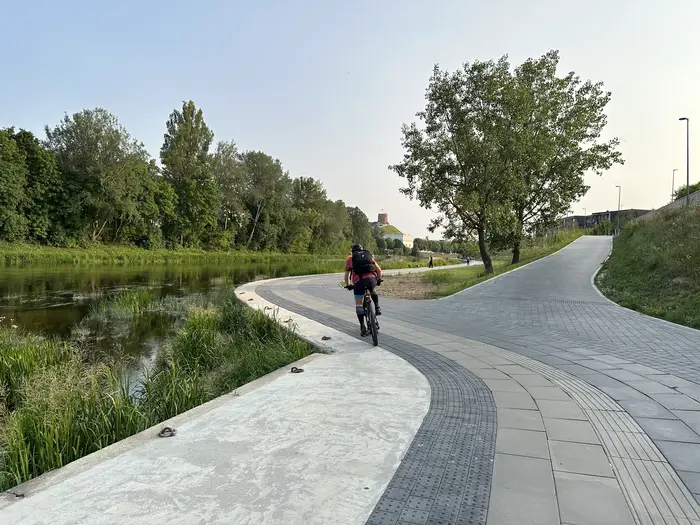 A cyclist riding on a paved riverside path next to the Neris River in Vilnius, with green trees and Gediminas' Tower on a hill in the background.