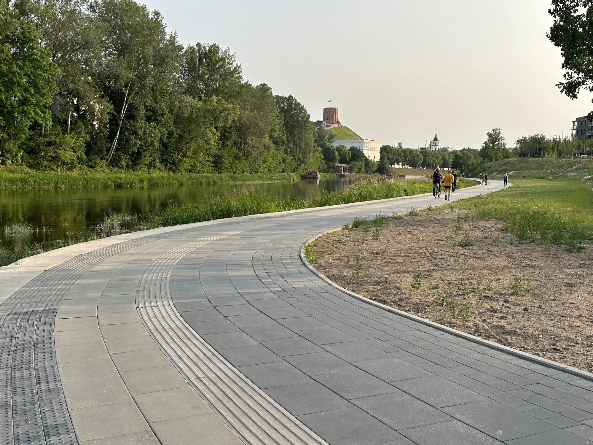 A curved grey stone pathway runs along the Neris River in Vilnius, with green trees on the opposite bank and Gediminas' Tower visible in the background.
