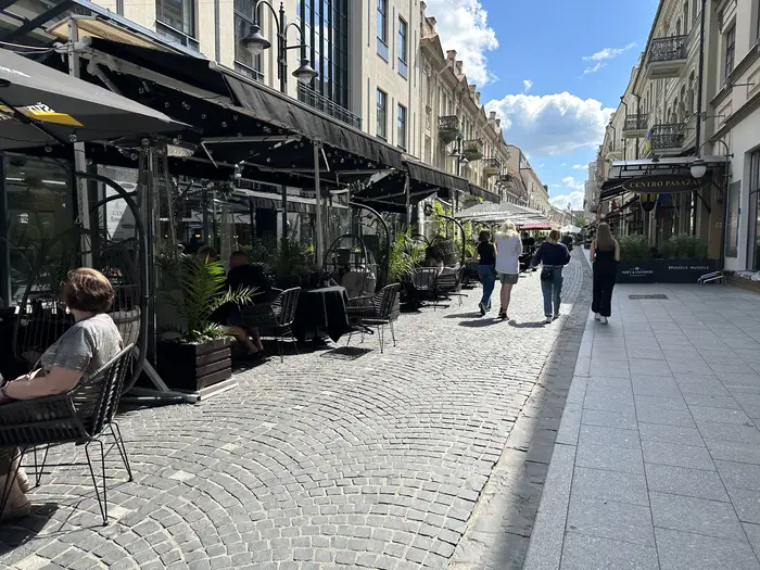 A cobblestone pedestrian street in Vilnius lined with outdoor cafe terraces under black awnings, with people walking under a blue sky.