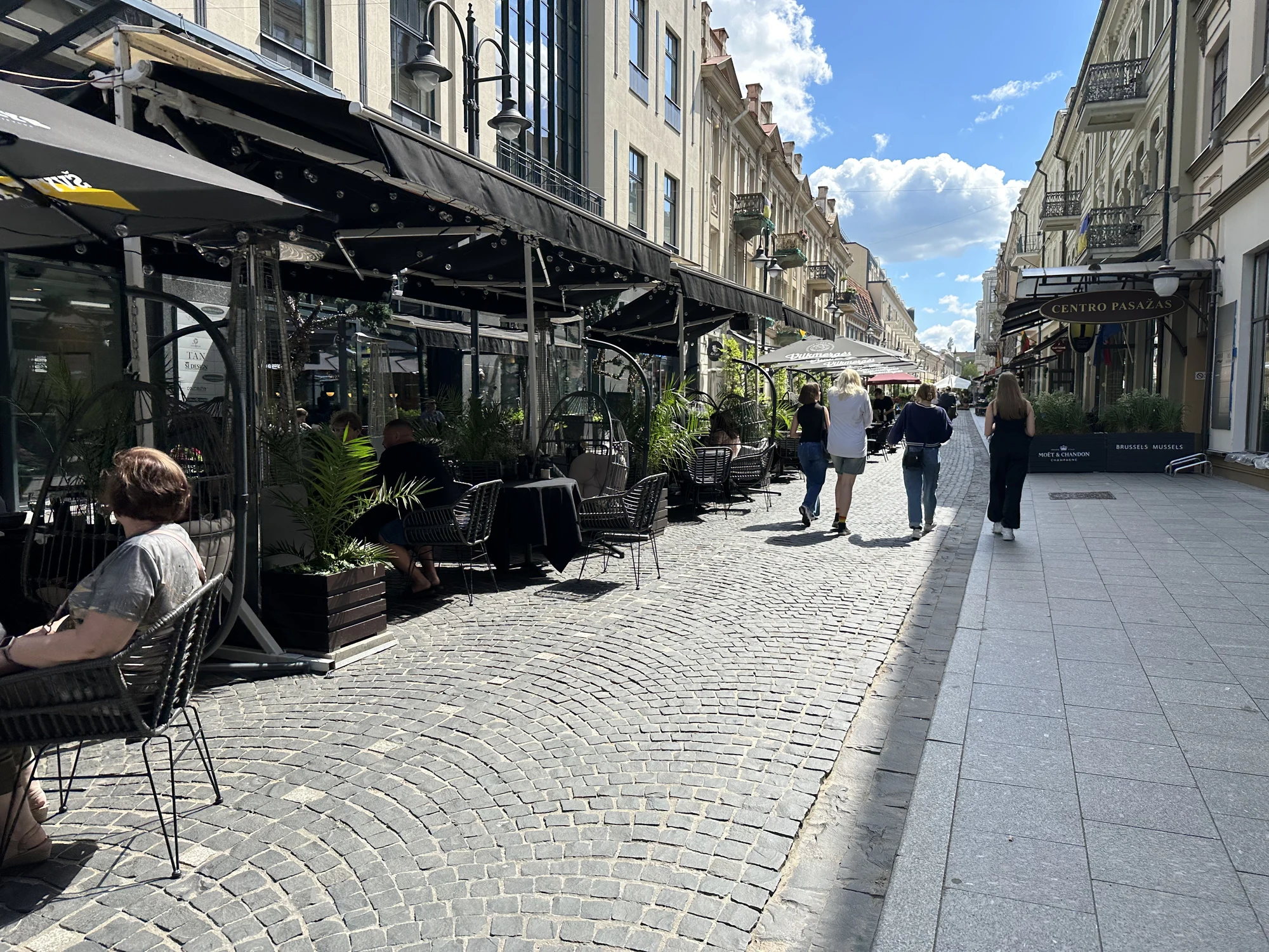 A cobblestone pedestrian street in Vilnius lined with outdoor cafe terraces under black awnings, with people walking under a blue sky.