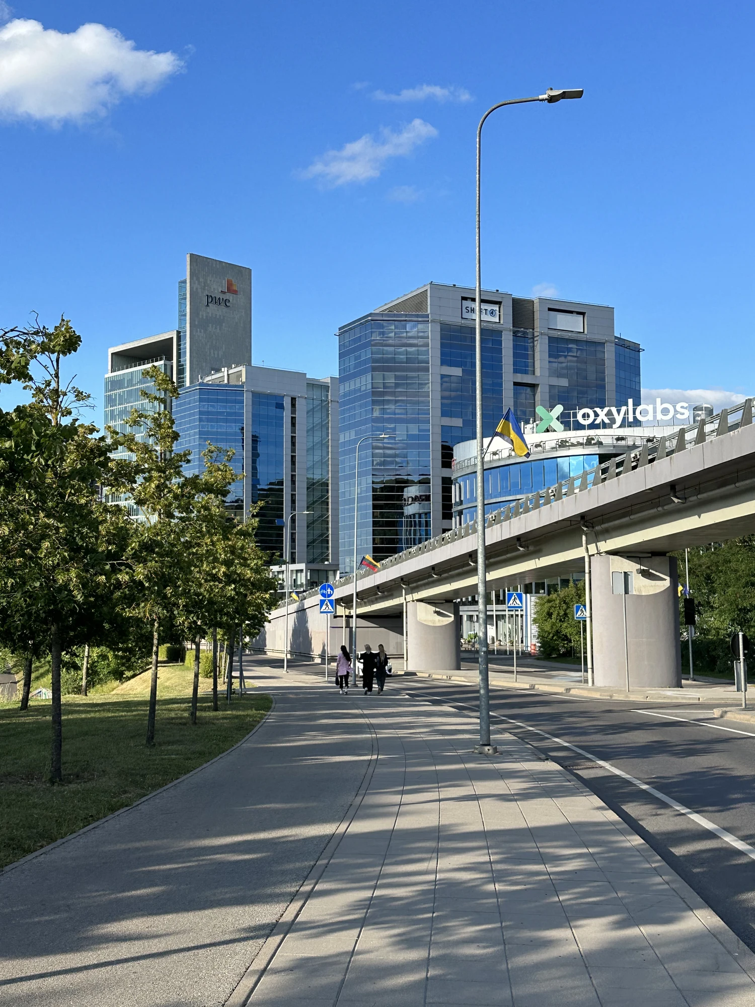 A paved pedestrian path leads towards modern glass office buildings with corporate logos under a bright blue sky in Vilnius.