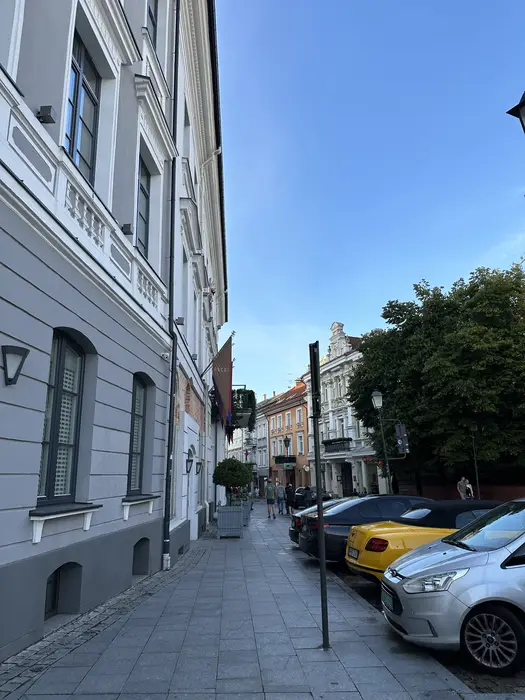 A sidewalk view along Didžioji Street in Vilnius Old Town, featuring the elegant grey facade of Hotel Pacai on the left and parked cars on the right under a clear blue sky.