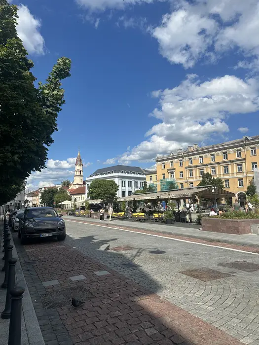 A cobblestone street in Vilnius Old Town featuring outdoor cafes with yellow chairs, historic buildings, a church spire in the background, and a parked car on the left under a blue sky.