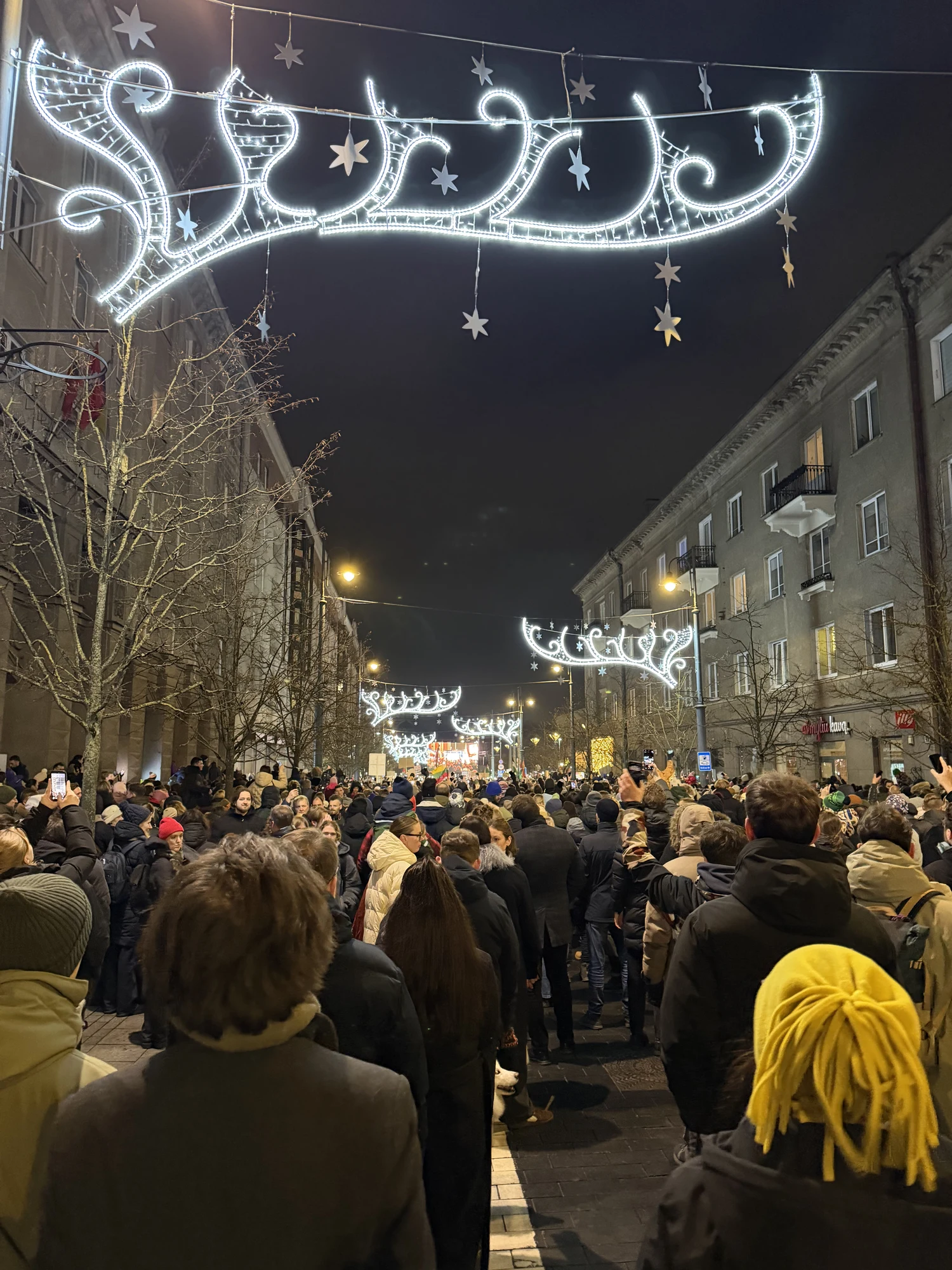 A view from behind a dense crowd of people walking down a city street at night, illuminated by bright, star-shaped holiday light arches hanging overhead.