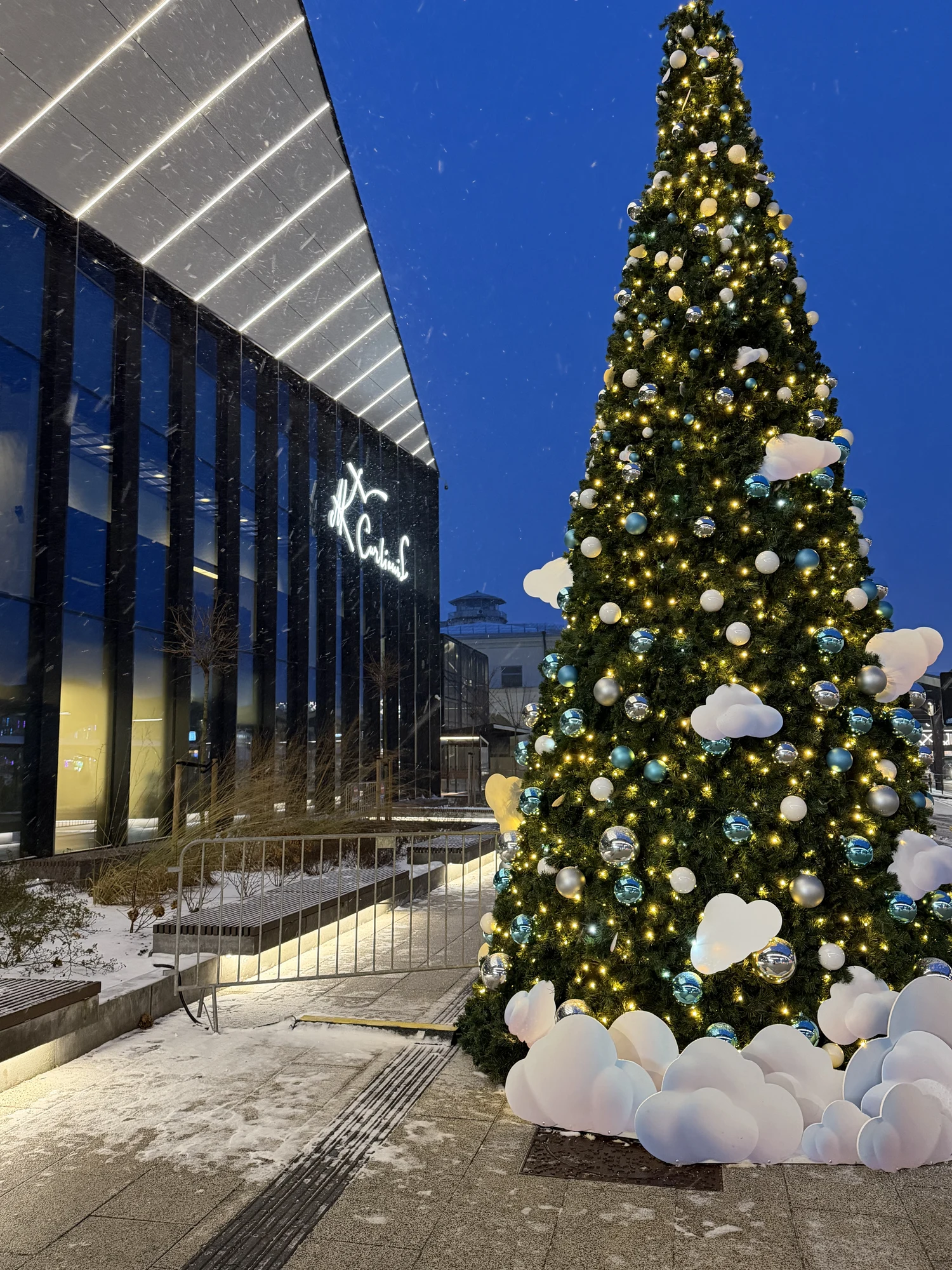A tall, illuminated Christmas tree decorated with blue and silver baubles and white cloud-shaped ornaments stands next to a modern glass-fronted building with the name 'M.K. Čiurlionis' lit up on its facade at dusk.