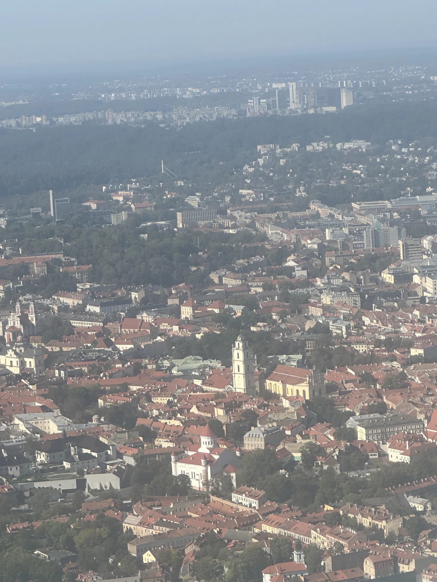 An aerial, slightly hazy view of Vilnius Old Town featuring historic red-roofed buildings, church spires, and distant modern skyscrapers under a clear sky.