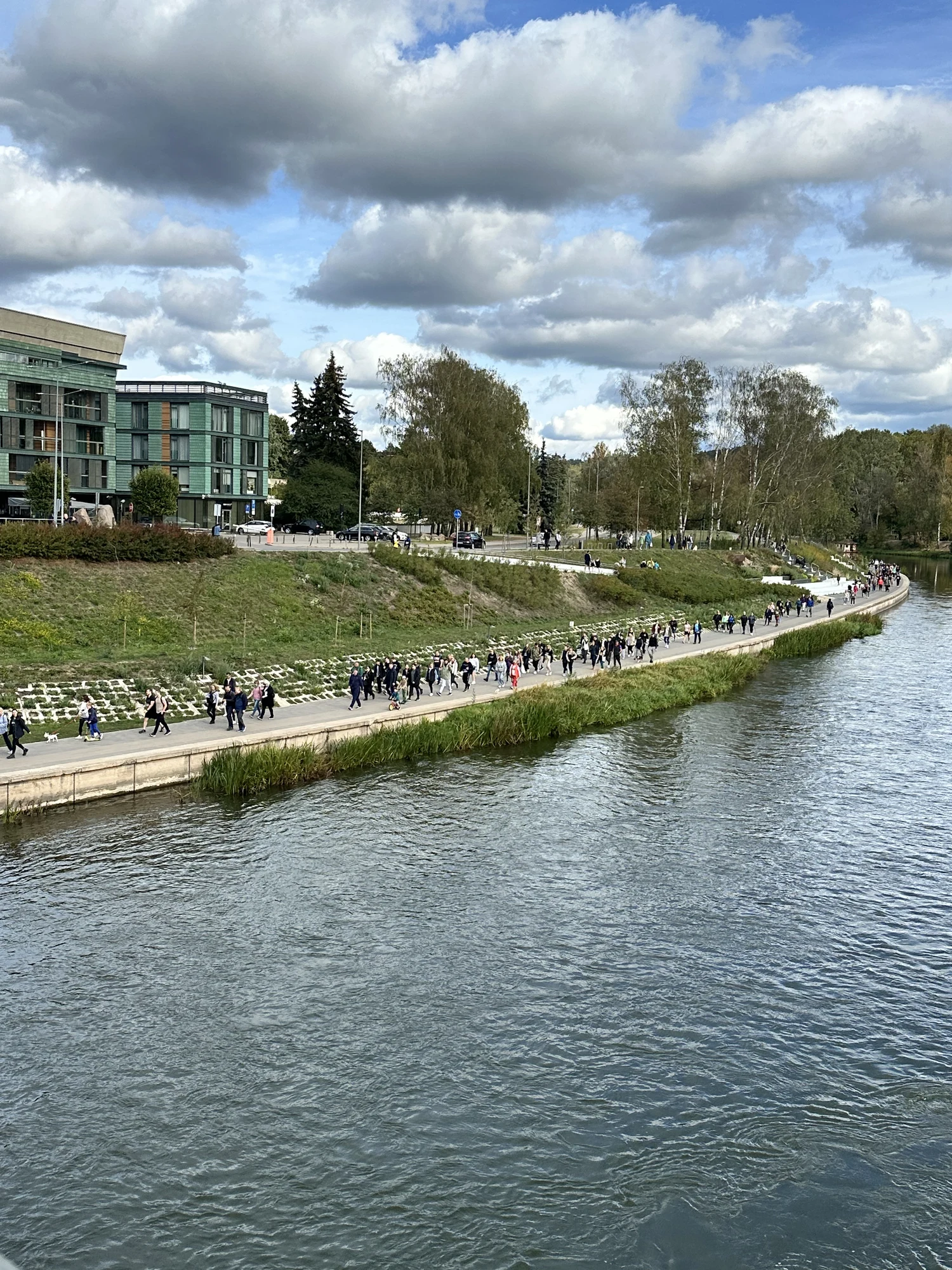 High-angle view of the Neris River in Vilnius with a crowded pedestrian path running along the green riverbank next to modern apartments.