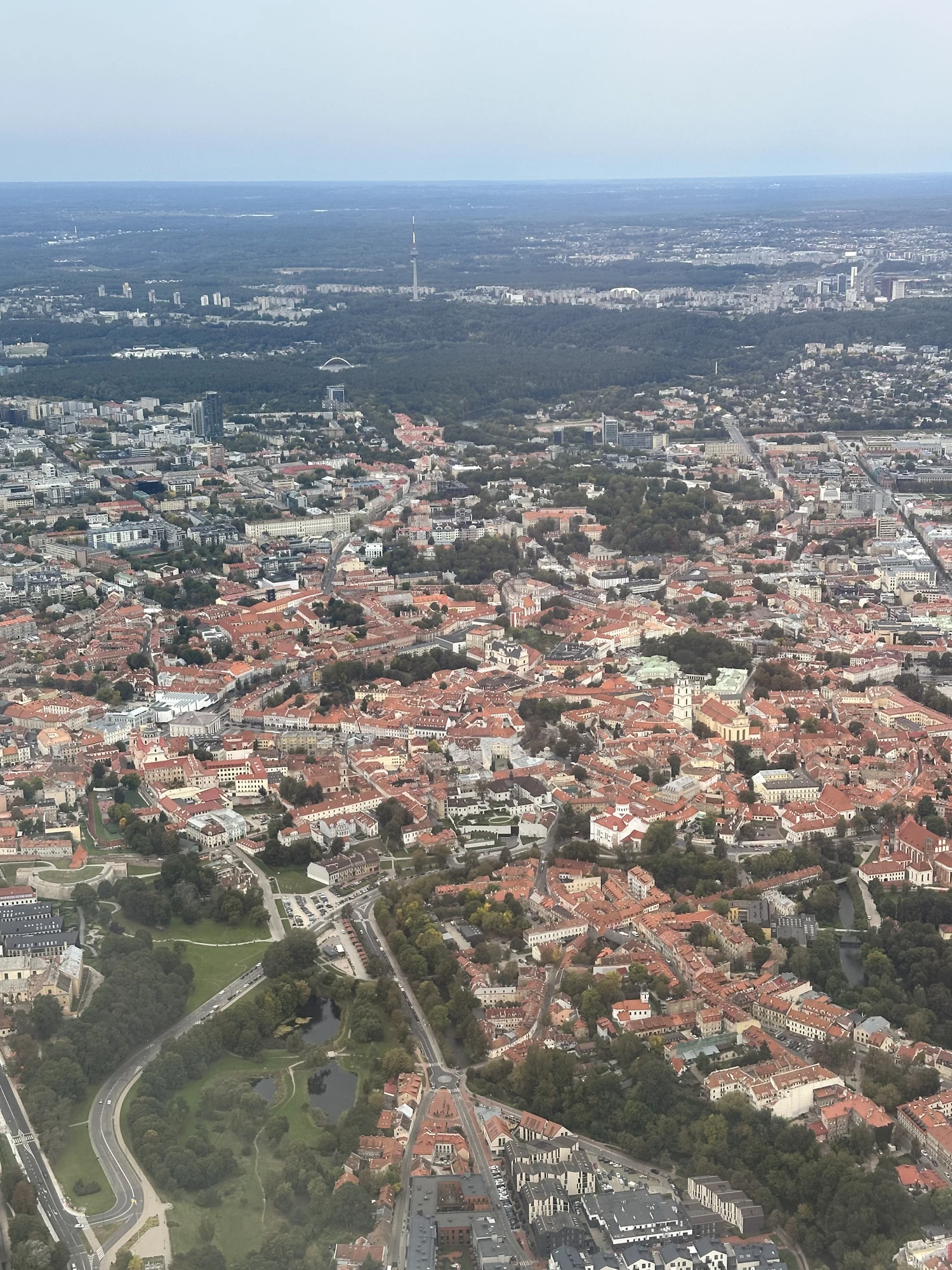 High-angle aerial view of Vilnius Old Town with its dense red-roofed buildings, green parks, and the TV tower visible in the far background under a hazy sky.
