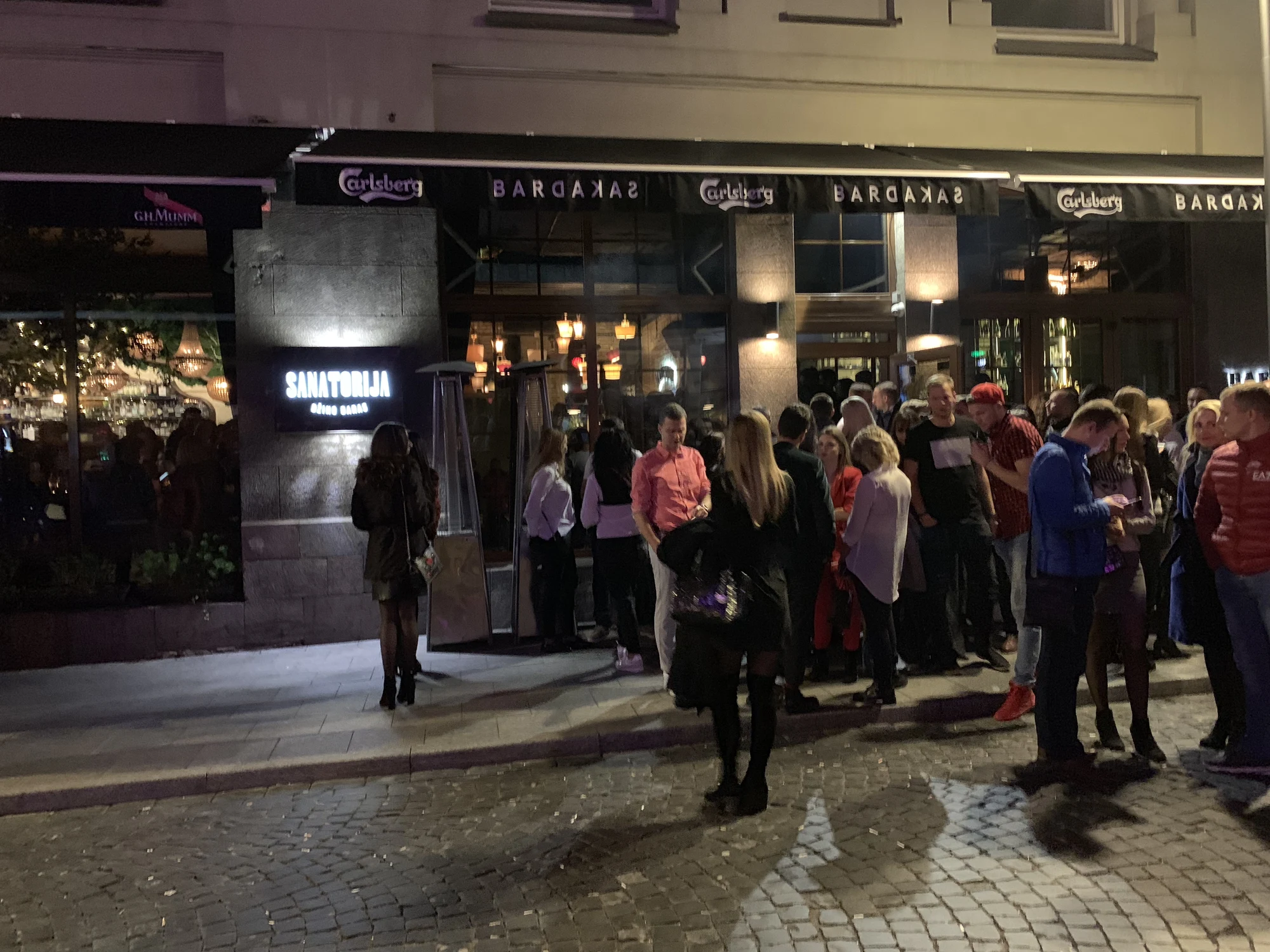 A crowd of people socializing on a cobblestone street outside the illuminated entrance of Sanatorija Gin Bar at night in Vilnius.