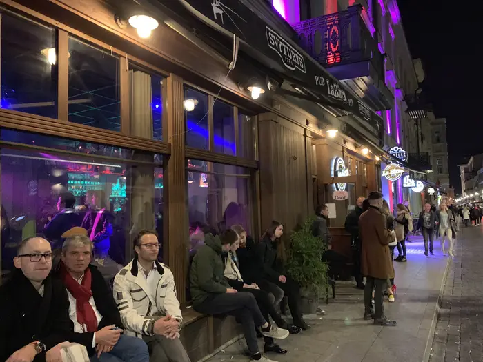 A group of people sitting on benches and standing outside La Birra Pub on a cobblestone street in Vilnius at night under purple and warm streetlights.