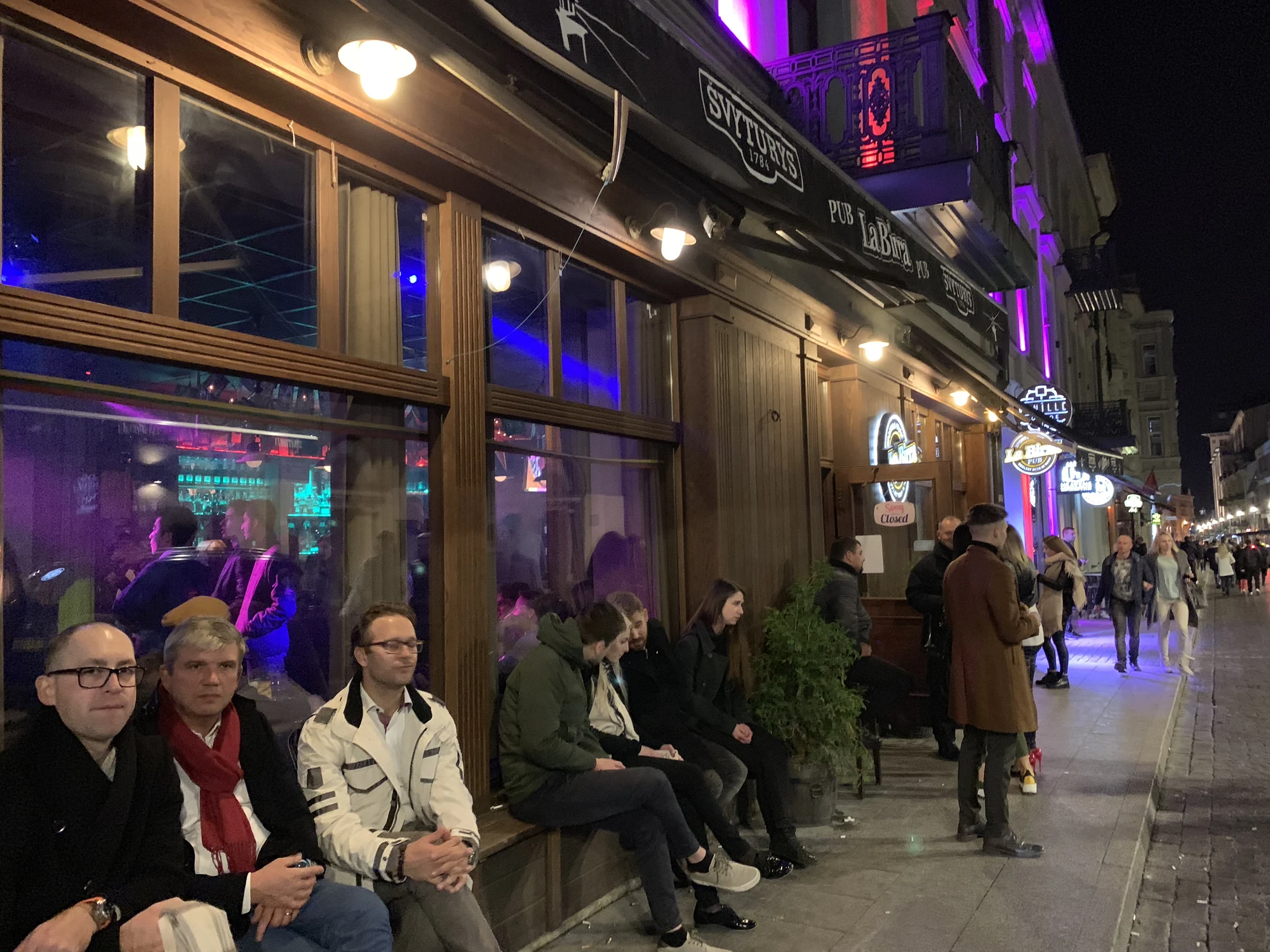 A group of people sitting on benches and standing outside La Birra Pub on a cobblestone street in Vilnius at night under purple and warm streetlights.