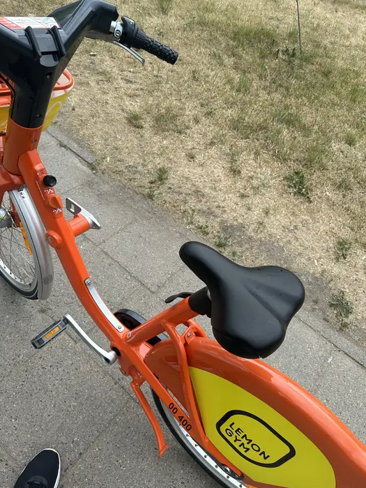 A close-up high-angle shot of an orange rental bicycle with a yellow mudguard featuring a Lemon Gym logo, parked on a paved sidewalk next to grass.