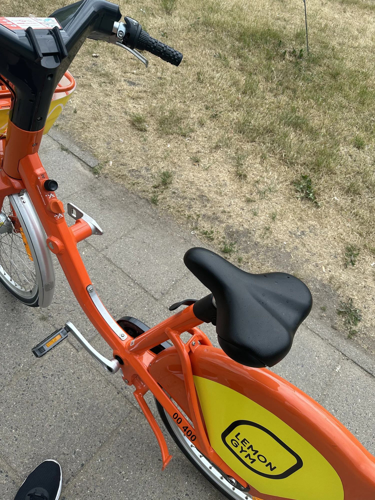 A close-up high-angle shot of an orange rental bicycle with a yellow mudguard featuring a Lemon Gym logo, parked on a paved sidewalk next to grass.