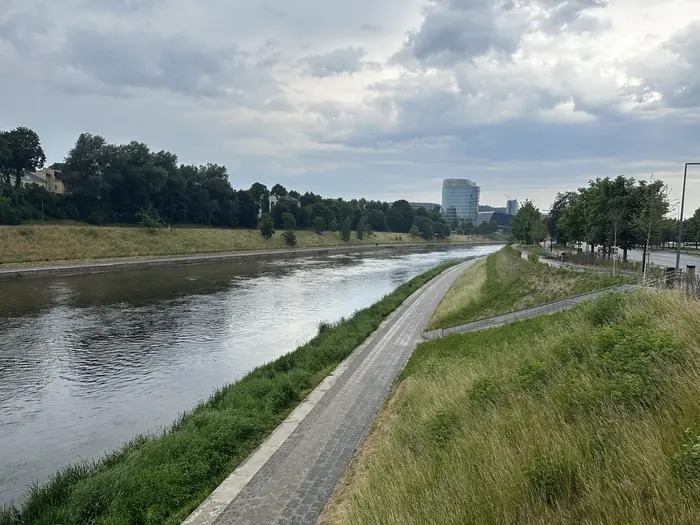 A paved pedestrian path runs alongside the Neris River in Vilnius, Lithuania, bordered by green grassy banks under an overcast sky.