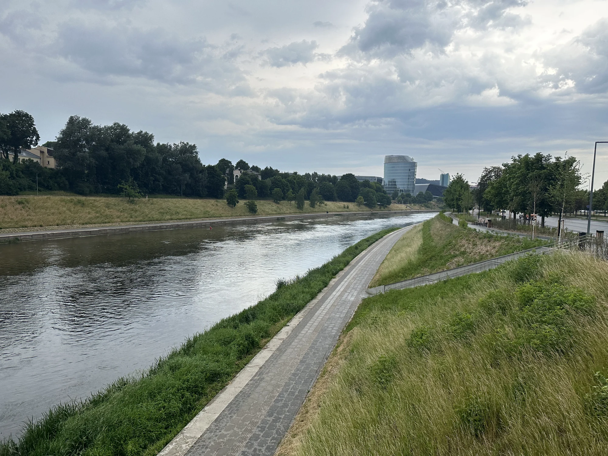 A paved pedestrian path runs alongside the Neris River in Vilnius, Lithuania, bordered by green grassy banks under an overcast sky.