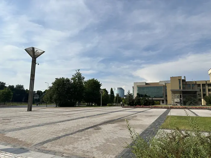 A wide paved public square in Vilnius with a tall, unique modernist street lamp on the left, lush green trees in the middle ground, and contemporary buildings under a soft blue sky.