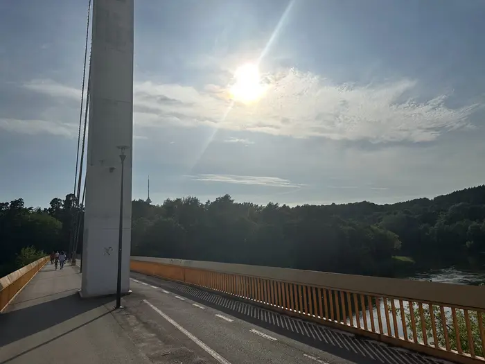 A wide shot of the pedestrian Vingis Park Bridge in Vilnius under a bright sun, with the Neris River and lush green forests in the background.