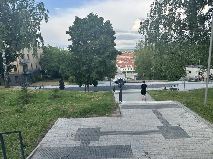 A paved stone pathway with stairs leading down a grassy hill towards trees, sculptures, and red-roofed buildings of Vilnius in the background under a cloudy sky.