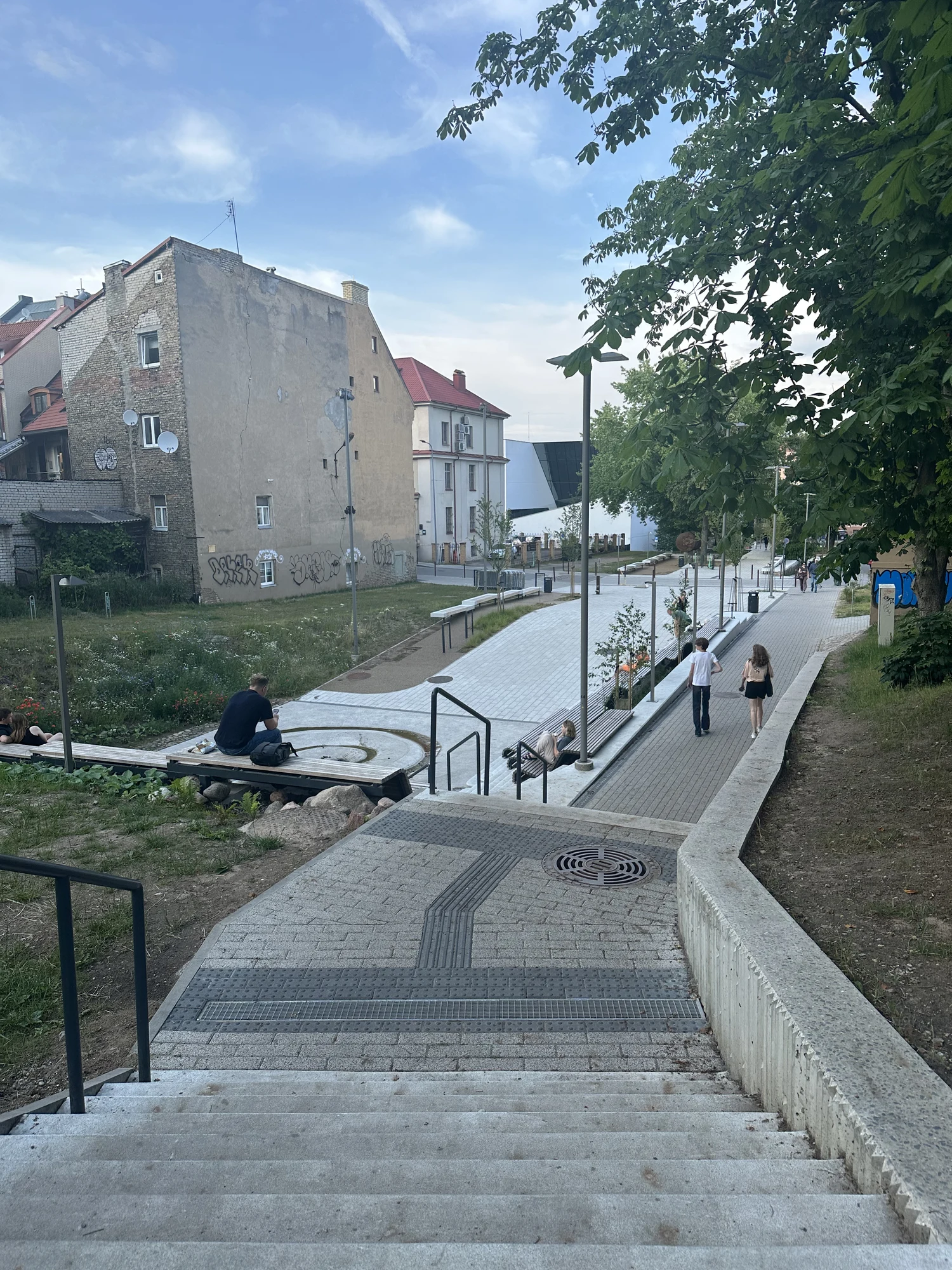 A view looking down concrete stairs into a modern paved public park with benches, green spaces, and historic buildings in Vilnius.