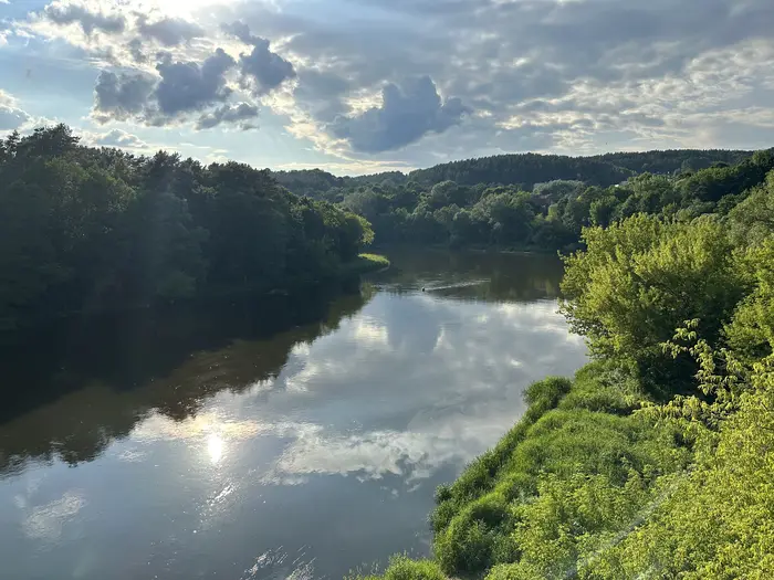 A wide view of the Neris River winding through dense green forests under a cloudy sky with sun reflecting on the water.