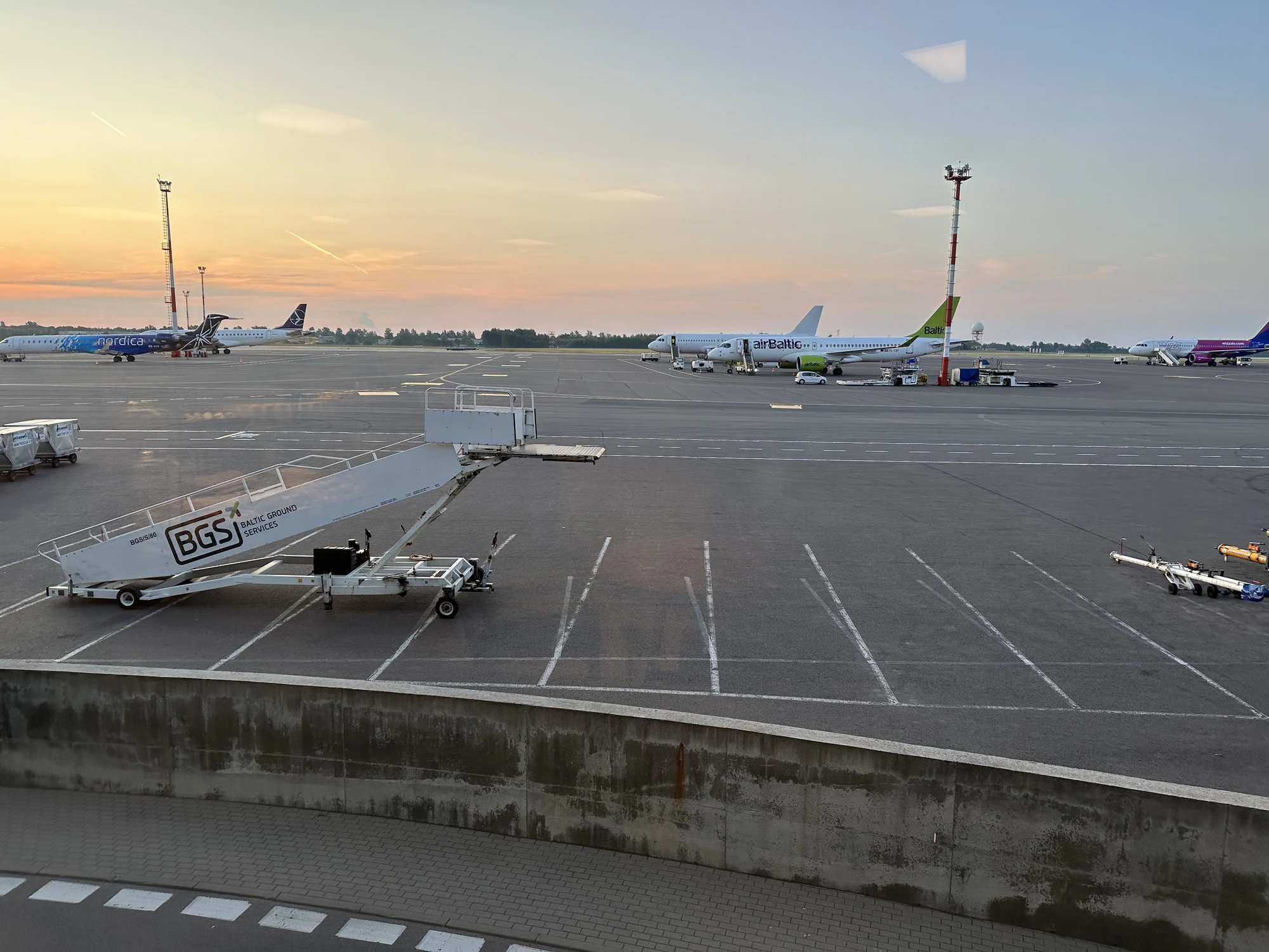 An airport apron at sunset with parked airplanes from airBaltic, Nordica, and Wizz Air, with mobile boarding stairs in the foreground.