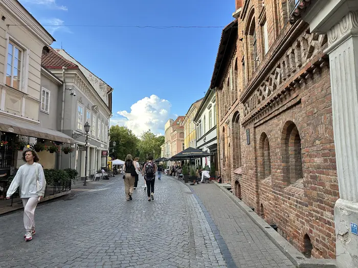 A cobblestone street in Vilnius Old Town with historic brick and plaster buildings, featuring people walking under a bright blue sky.
