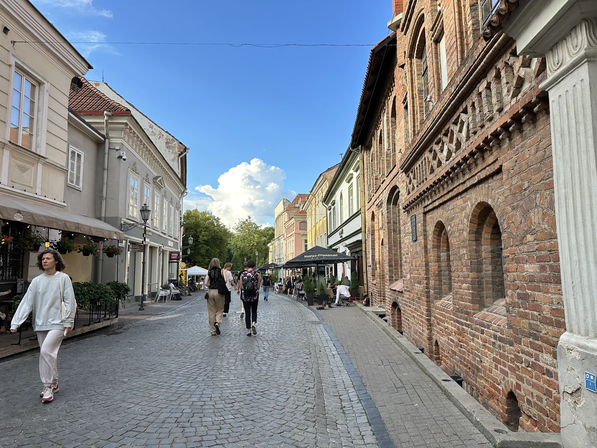 A cobblestone street in Vilnius Old Town with historic brick and plaster buildings, featuring people walking under a bright blue sky.