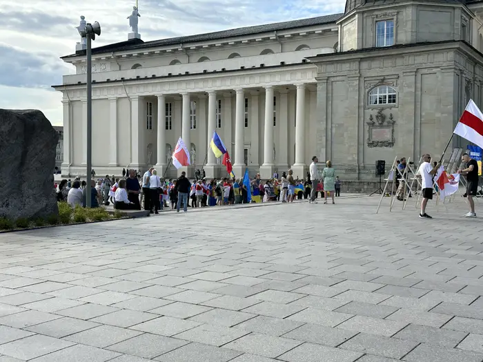 A crowd of people holding flags, including Ukrainian and Belarusian opposition flags, gathered in the paved Cathedral Square in front of the neoclassical Vilnius Cathedral.