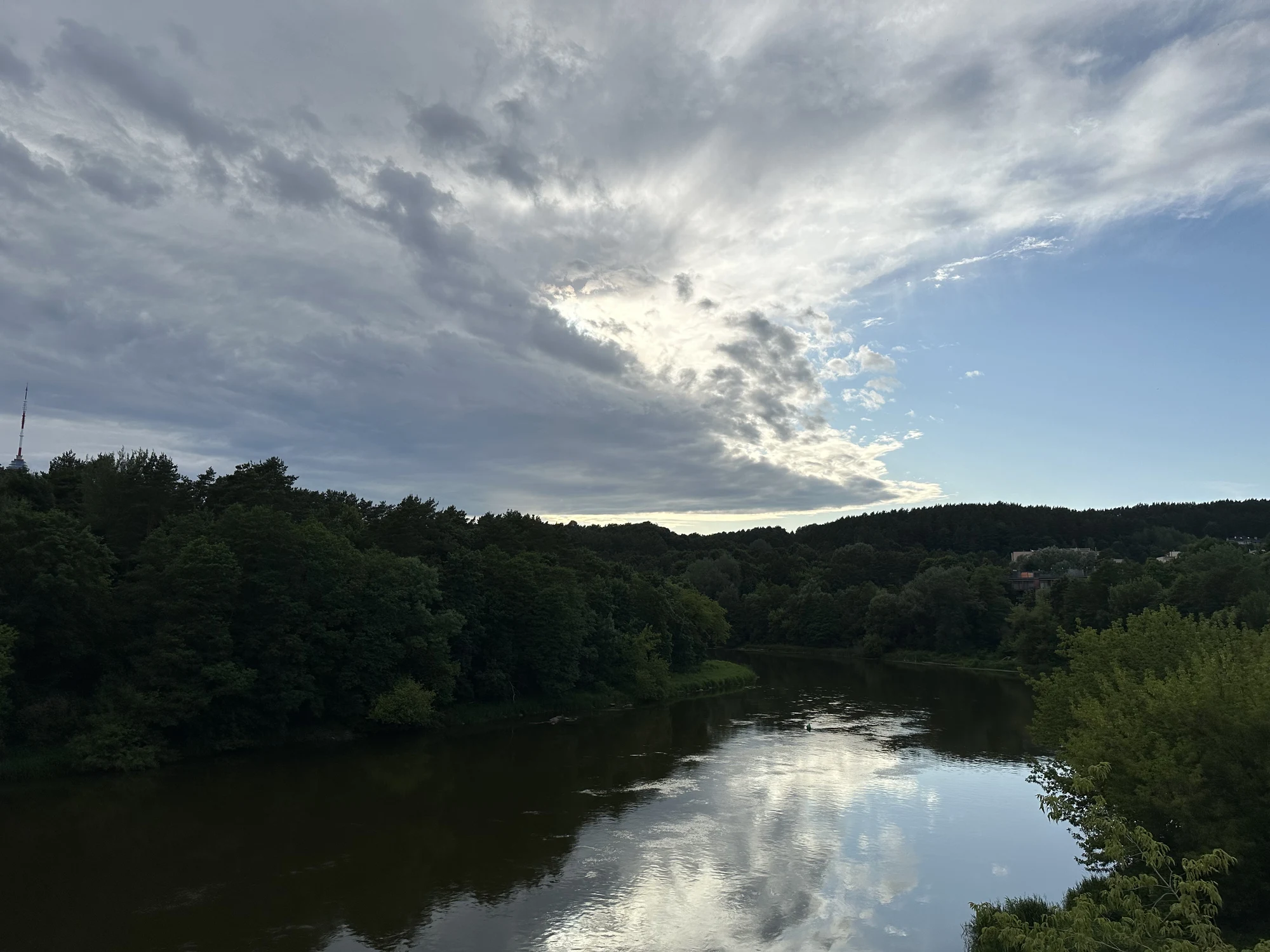 A wide shot of the Neris River in Vilnius surrounded by dense green forests under a cloudy sky, with the Vilnius TV Tower visible in the far distance.