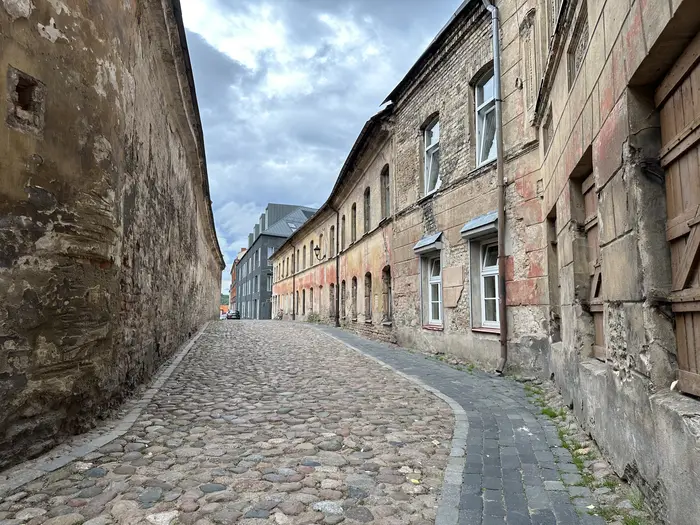 A curving cobblestone street in Vilnius Old Town flanked by historic, weathered buildings under a cloudy sky.