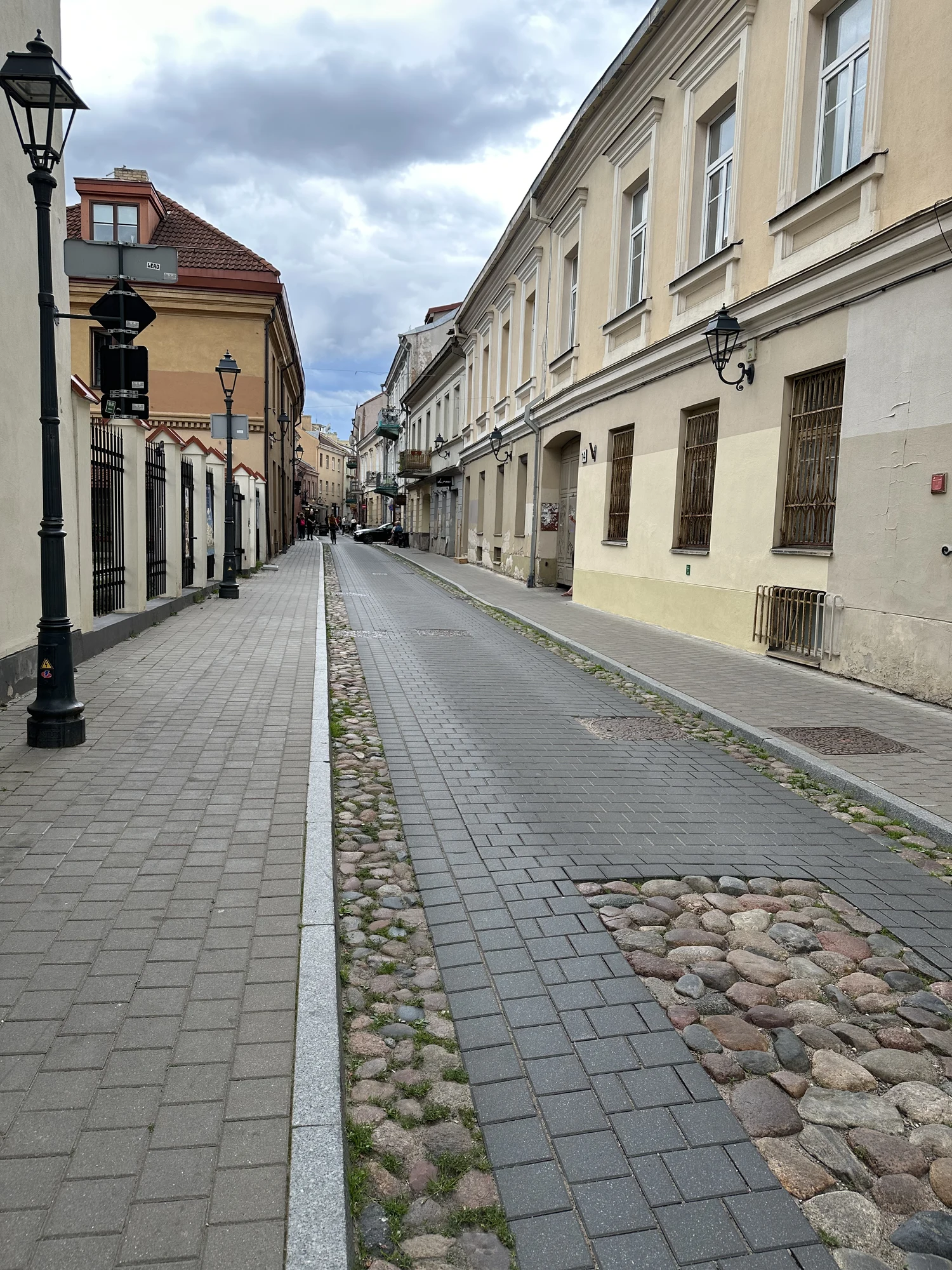 A narrow paved street in Vilnius Old Town with historic buildings, classic black street lamps, and sections of exposed cobblestones under an overcast sky.