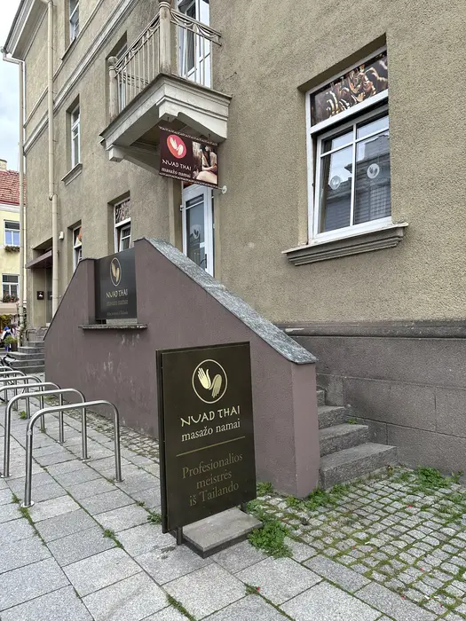 A street-level view of the Nuad Thai massage parlor entrance in Vilnius, featuring brown signs with gold lettering and a small staircase leading to the door.