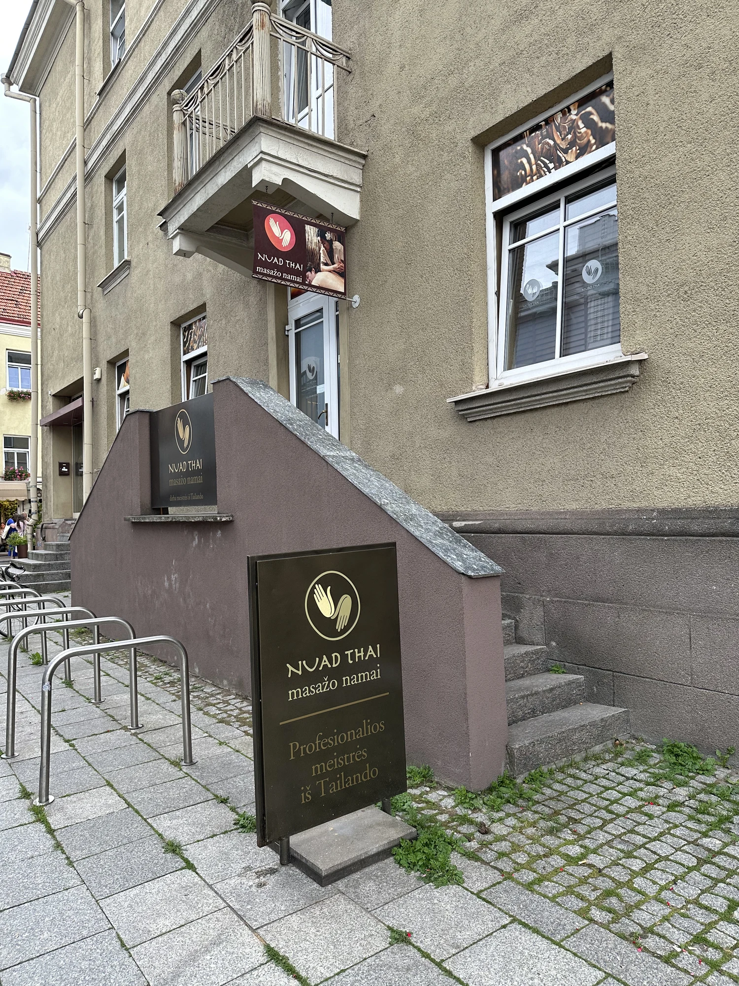 A street-level view of the Nuad Thai massage parlor entrance in Vilnius, featuring brown signs with gold lettering and a small staircase leading to the door.