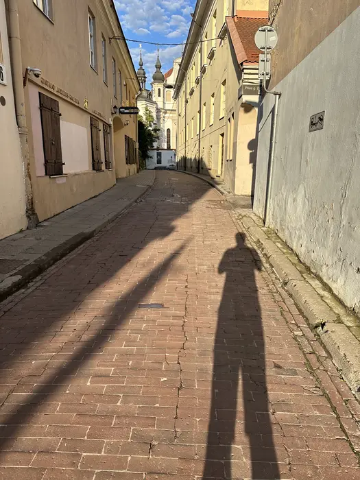 A vertical view down a narrow cobblestone street in Vilnius Old Town, featuring the long shadow of the photographer in the foreground and church towers in the background.