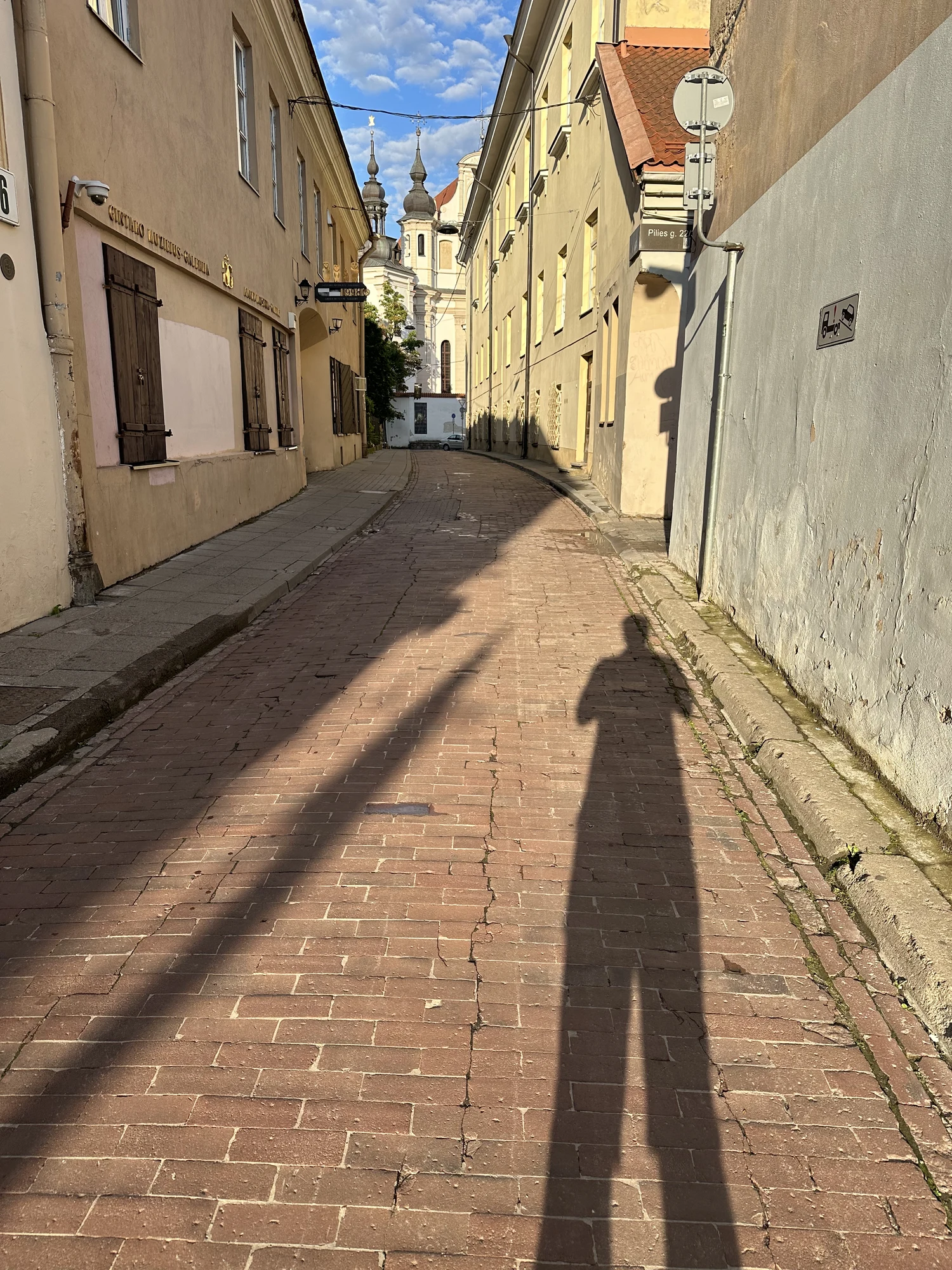 A vertical view down a narrow cobblestone street in Vilnius Old Town, featuring the long shadow of the photographer in the foreground and church towers in the background.