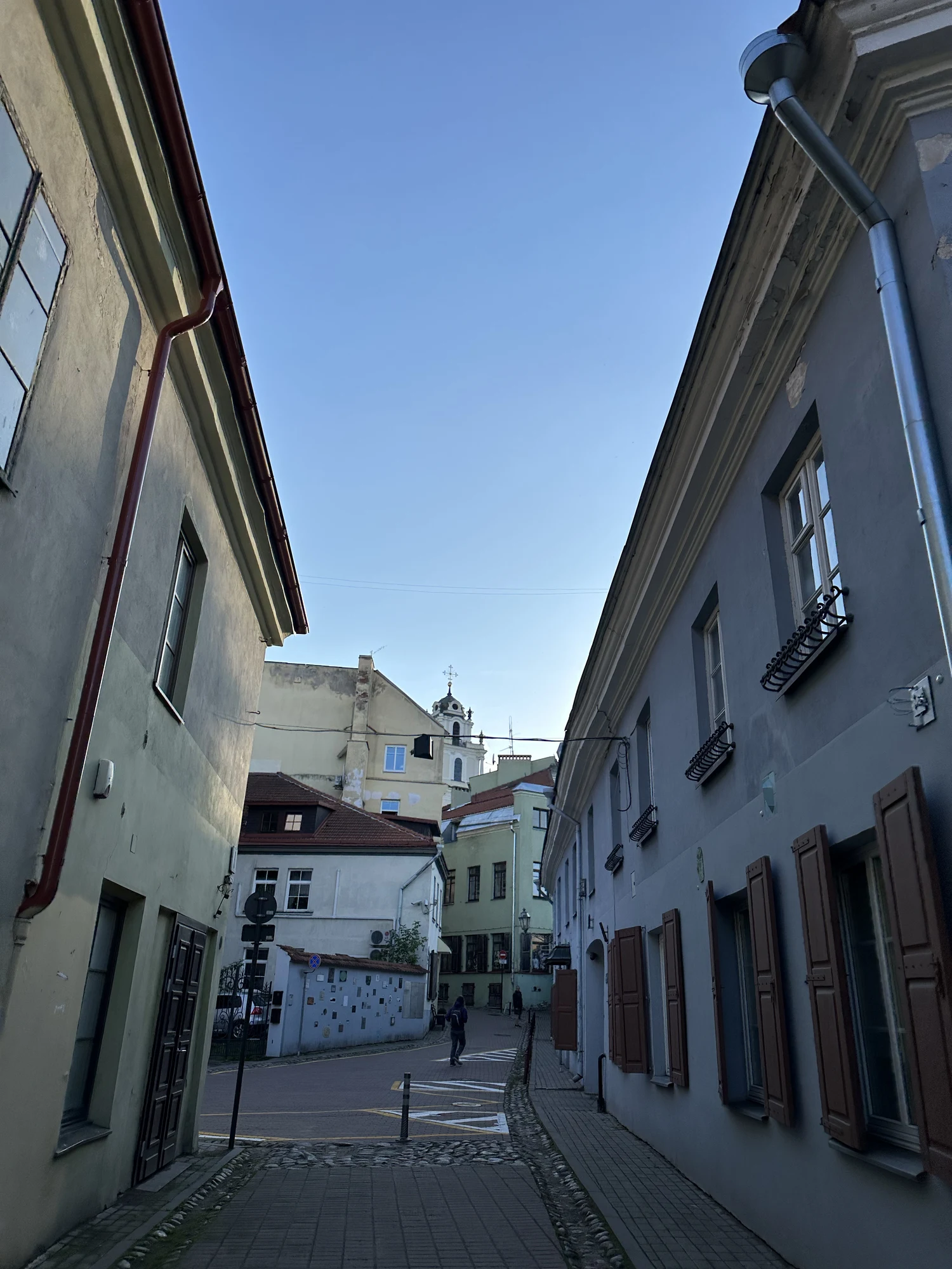 A narrow cobblestone street in Vilnius Old Town lined with historic pastel-colored buildings, leading towards a baroque church tower under a clear blue sky.