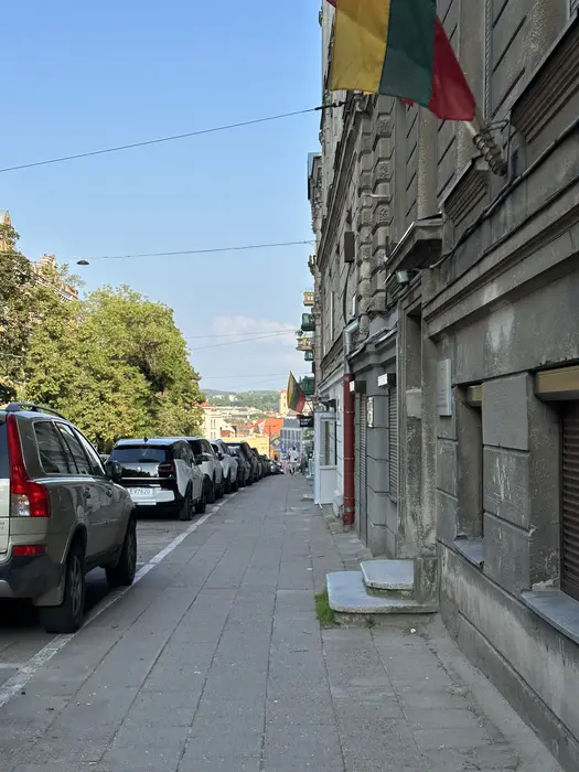 A paved sidewalk in Vilnius with a stone building on the right displaying a Lithuanian flag, and a row of parked cars on the left under a clear blue sky.