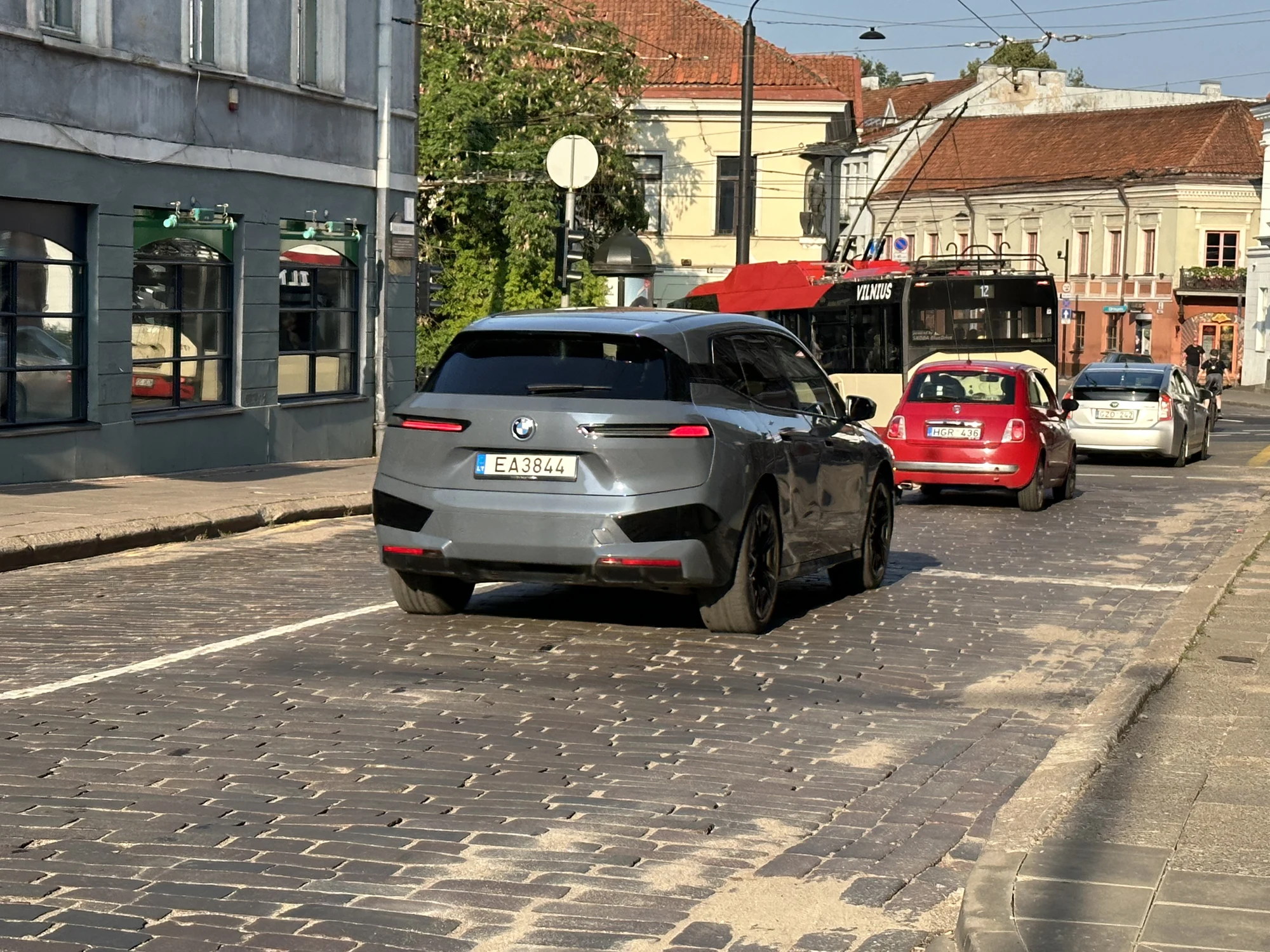 A grey BMW SUV drives on a cobblestone street in Vilnius, Lithuania, with a red trolleybus and other cars in the background.