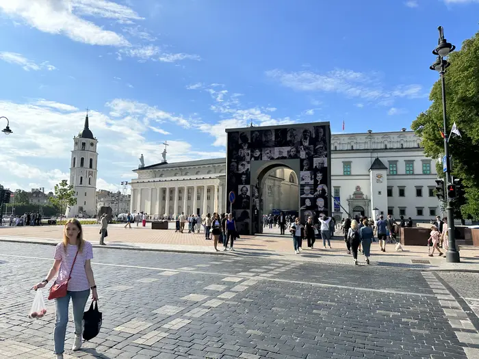 A wide shot of Cathedral Square in Vilnius, Lithuania, showing the white cathedral, its detached bell tower, the Palace of the Grand Dukes, and people walking on the cobblestone plaza.