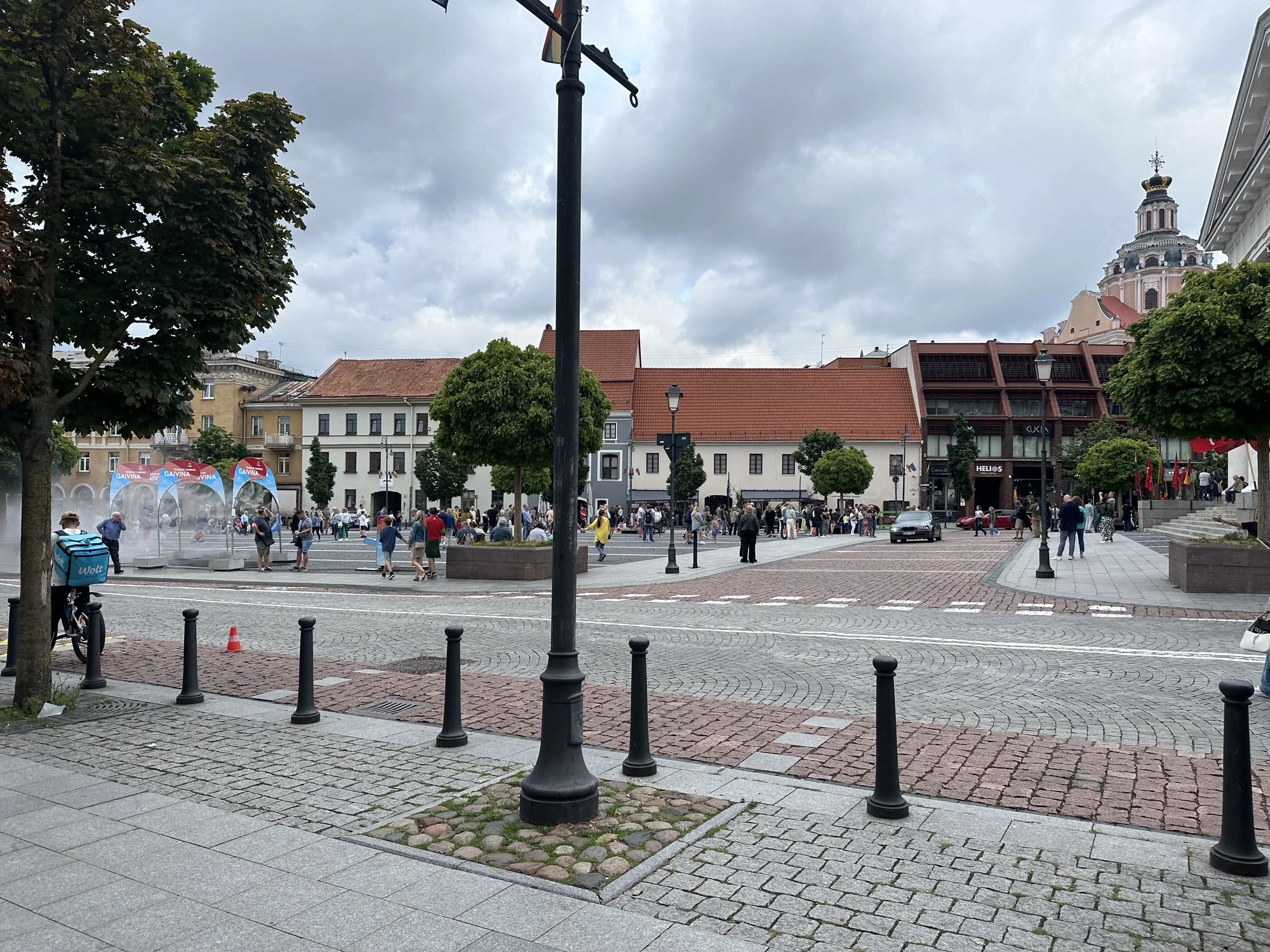 A wide view of Town Hall Square in Vilnius, featuring cobblestone paving, historic buildings, people walking, and the dome of St. Casimir's Church in the background under a cloudy sky.