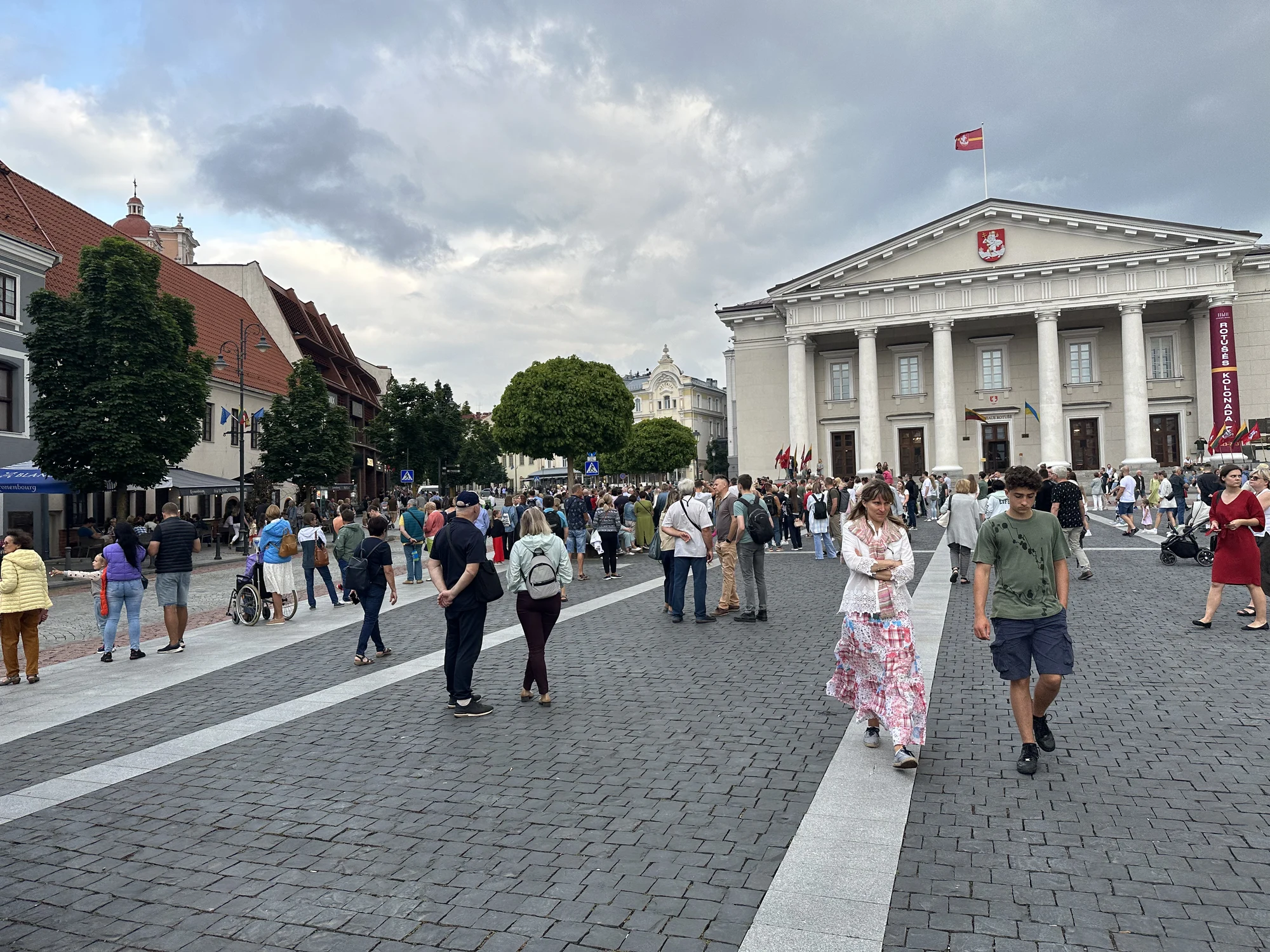 A bustling cobblestone square in front of the grand neoclassical Vilnius Town Hall, filled with people walking under an overcast sky.