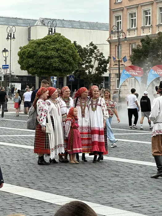 A group of five women and one young girl in traditional red and white Lithuanian folk dresses and headscarves standing on the cobblestones of Vilnius Town Hall Square.